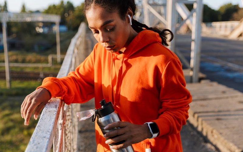 african-american-serious-woman-sportswear-using-earpods-holding-water-bottle-while-leaning-railing-old-bridge-min